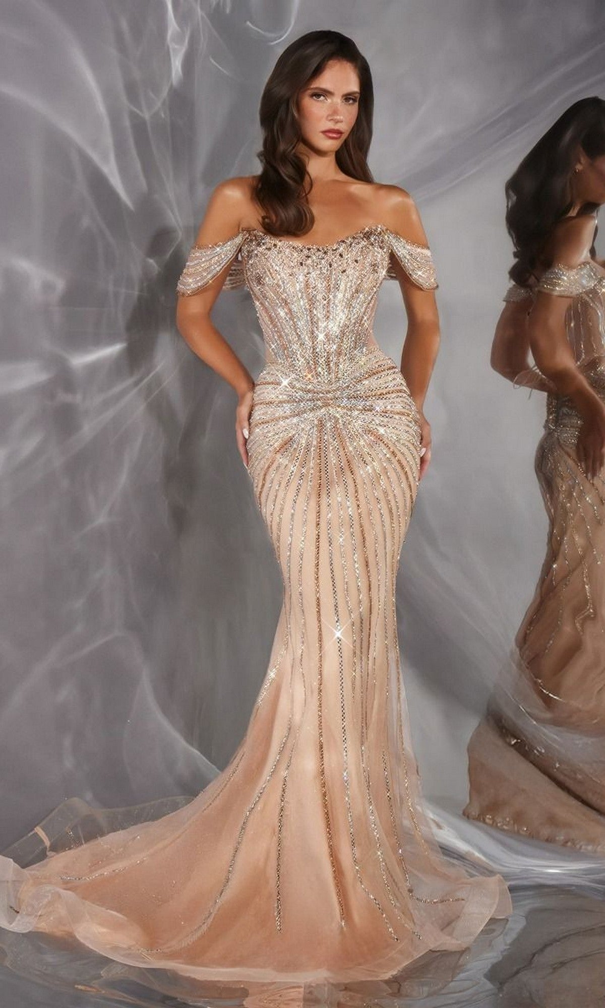 A woman wearing the Ladivine CD654 Long Prom Dress in gold, an off-the-shoulder gown with a fitted, floor-length silhouette adorned with sparkling crystal embellishments, stands in front of a gray textured backdrop with her reflection visible in a mirror beside her.